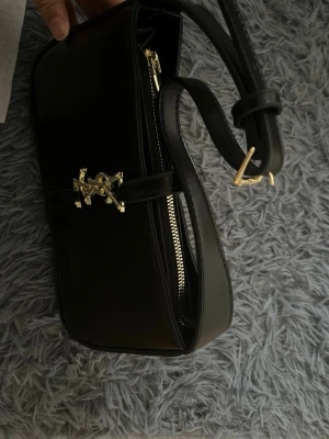 Svart axelväska från Saint Laurent - Säljer en stilren svart axelväska från Saint Laurent i slätt skinn med justerbar rem och guldfärgad YSL-logga framtill. Väskan har en elegant form och stängs med dragkedja. Perfekt för dig som gillar klassiska designer och snygga detaljer.