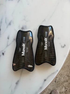 Monolit benskydd i kolfiber - Säljer ett par svarta benskydd från Monolit i kolfiber med snyggt mönster och vit logga. De är lätta och har en ergonomisk form som passar bra för fotboll. Perfekt för dig som vill ha skydd med stil på planen.