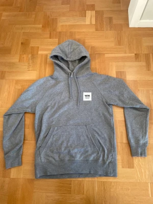 Wood Wood Hoodie - En grå Wood Wood Hoodie i mycket bra skick. Enda synliga deffekterna är lite slitningar på loggorna, se bild. Storlek: Small