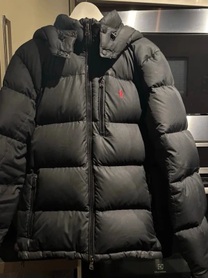 Svart Polo Ralph Lauren	Dunjacka   - Svart dunjacka från Polo Ralph Lauren med röd broderad logga på bröstet. Jackan har hög krage, dragkedja framtill och flera fickor med dragkedja. Materialet är polyester och dun, perfekt för kalla dagar. Insidan har ett diskret rutigt mönster.