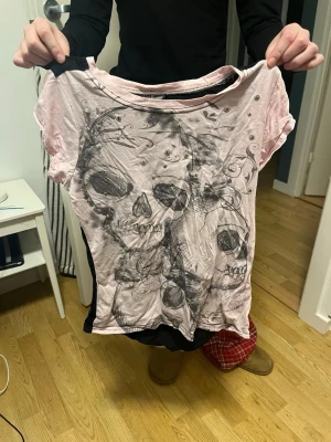 Rosa och svart t-shirt med döskallar - Cool t-shirt i rosa och svart med stort döskalletryck framtill och små pärldetaljer. Framsidan har konstnärligt motiv med flera döskallar och dekorativa mönster, medan baksidan är helsvart. Rund hals och korta ärmar, perfekt för dig som gillar edgy stil.