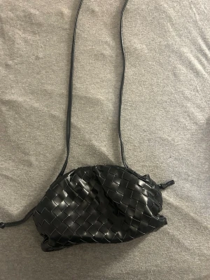Svart flätad axelväska i skinn - Säljer en svart axelväska med lång rem och snyggt flätad design. Väskan är gjord i skinn och har en praktisk öppning med snörning upptill. Aldrig använd 