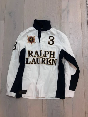 Vit och svart pikétröja Ralph Lauren - Snygg långärmad pikétröja från Ralph Lauren i vit och svart färg. Stor broderad logga och siffran 3 på både fram- och baksidan samt på ärmen. Tröjan har ståkrage med knappar och detaljer i guld. Perfekt för dig som gillar sportig och klassisk stil.