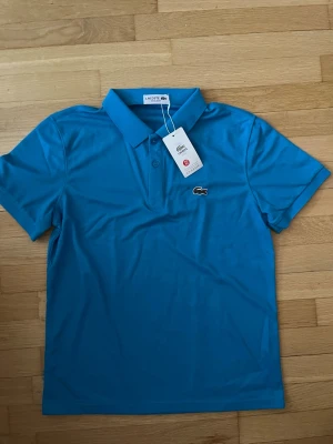 Blå pikétröja från Lacoste M - Säljer en klassisk blå pikétröja från Lacoste i storlek M. Tröjan har krage, två knappar framtill och den ikoniska krokodilloggan broderad på bröstet. Materialet är mjukt och ventilerande, perfekt för en chill och sportig look.