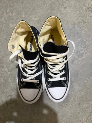 Svarta Converse All Star high tops - Säljer ett par klassiska svarta Converse All Star high tops med vit tå och vita snören. Skorna har rund tå, platt sula och tydliga vita sömmar. Märket syns på baksidan och insidan. Materialet är bomullstyg med gummisula. Perfekt för dig som gillar ikoniska sneakers.