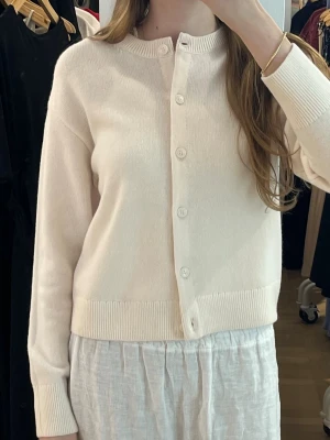 Vit kofta med knappar - Säljer en stilren vit kofta med rund hals och knäppning framtill. Koftan har långa ärmar och ribbade muddar vid ärmslut och nederkant. Perfekt att slänga på sig när det är lite kyligt ute. Enkel och clean design som passar till det mesta. 100% merino ull.