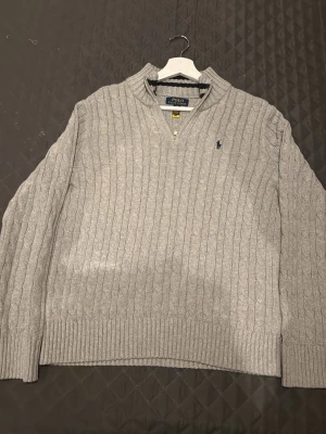 Grå stickad tröja Polo Ralph Lauren - Säljer en grå kabelstickad tröja från Polo Ralph Lauren med half zip och långärmad design. Tröjan har ribbade muddar och krage, samt en liten broderad logga på bröstet. Perfekt för dig som gillar klassisk och stilren look.