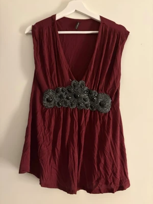 Vinröd blus med pärldetaljer från Amisu - Säljer en vinröd blus från Amisu i storlek L. Toppen har djup v-ringning och är ärmlös. Midjan är dekorerad med svarta pärlor och broderade detaljer som ger en unik look. Materialet är mjukt och lätt, perfekt för att sticka ut.