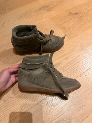 Isabel Marant wedge sneakers i grön mocka - Säljer ett par Isabel Marant wedge sneakers i grön mocka med bruna gummisulor. Skorna har snörning, perforerade detaljer och en dold kilklack som ger extra höjd. Snygga vågiga kanter och rund tå. Perfekta för dig som gillar coola och trendiga sneakers. Litet märke längst fram annars i jättefint skick!