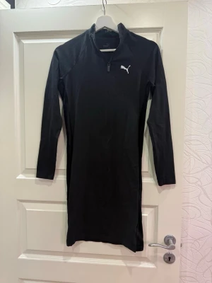 Svart långärmad klänning från Puma - Svart långärmad klänning från Puma med vit logga på bröstet. Klänningen har en halv dragkedja framtill och är tillverkad i mjuk bomull. Modellen är rak och har en liten slits på sidan. Storleken passar mer till xs/s