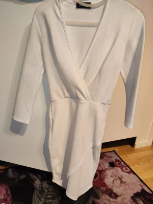 Vit omlottklänning med v-ringning - Säljer en snygg vit omlottklänning från Chiara Forthi med v-ringad hals och långärmad design. Klänningen har en kort längd och en asymmetrisk nederdel som ger en modern look. Materialet är mjukt och stretchigt, perfekt för dig som gillar stilrena plagg.