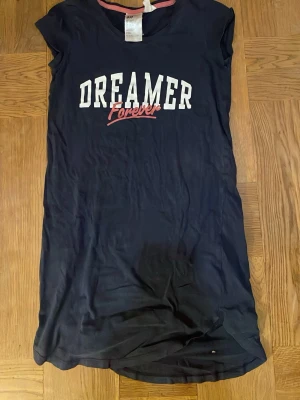 Marinblå nattklänning Dreamer Forever H&M - Snygg marinblå nattklänning från H&M med trycket 'Dreamer Forever' i vitt och rött på bröstet. Klänningen har rund hals och är kortärmad, tillverkad i mjuk bomull. Perfekt för dig som gillar avslappnad och cool stil. Väl använd i bra skick.