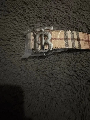 Rutig beige skärp med B-spänne - Säljer ett klassiskt Burberry-inspirerat skärp i beige med rutigt mönster och silverfärgat B-spänne. Skärpet är gjort i skinnimitation och har detaljer med ingraverade hästar. Perfekt accessoar för att lyfta din outfit.