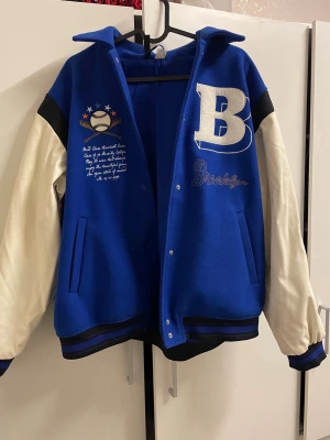 Blå H&M bomberjacka med Brooklyn-tryck - Snygg blå bomberjacka med vita ärmar och stora broderade detaljer som ett B och Brooklyn på bröstet. Jackan har tryckknappar, ribbade muddar och coolt basebollmotiv. Perfekt för dig som vill sticka ut med en sportig och trendig look.
