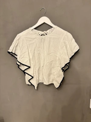 Vit blus med volangärmar och svart kant - Säljer en vit blus med korta volangärmar och svart kant längs ärmsluten. Toppen har en rund halsringning och knytning i nacken som ger en söt detalj. Vet ej storlek men skulle säga XS.