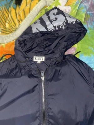 Svart vindjacka från Risky XS - Svart vindjacka från Risky med coolt tryck på huvan och ärmen. Jackan har hel dragkedja framtill, huva med snörning och är perfekt för dig som gillar streetwear och vill ha en snygg jacka med attityd. Helt ny oanvänt. 