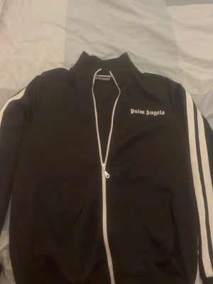 Svart Palm Angels zipper M - Palm Angels svart zipper med vita ränder längs ärmarna och vit dragkedja. Klassisk hög krage och logga på bröstet. Jackan har en slim passform och är tillverkad i polyester för en sportig vibe.
