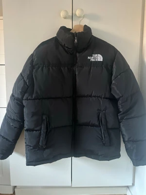 Svart pufferjacka från The North Face - Säljer en svart pufferjacka från The North Face med hög krage och synligt logotryck på bröstet och ryggen. Jackan har två fickor med dragkedja och en quiltad design som ger en schysst volym. Perfekt för kalla dagar och riktigt snygg till streetwear.