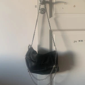 Svart axelväska med kedjedetaljer - Cool svart axelväska i skinnimitation med grova silverfärgade kedjor och snygga snörningsdetaljer längs kanterna. Väskan har ett stort fack och en axelrem med kedja som ger en edgy vibe. Perfekt för dig som gillar att sticka ut med accessoarer.
