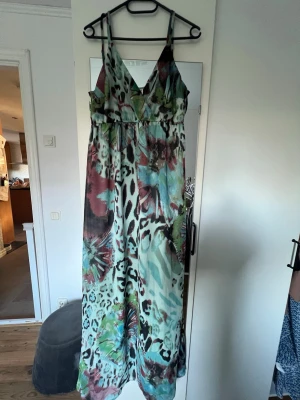 Mönstrad långklänning Vero Moda - Säljer en färgstark långklänning från Vero Moda i storlek L. Klänningen har ett vackert mönster med inslag av turkos, röd, svart och grön, samt leopardliknande detaljer. Den har v-ringad hals, smala justerbara axelband och är ärmlös. Ena axelbandet har gått av och knutits, se bild. I övrigt väldigt fin.
