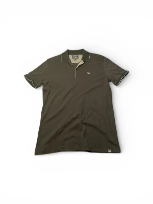 Svart piké från Woolrich - Svart pikétröja från Woolrich med klassisk krage och knappslå framtill. Tröjan har korta ärmar med ljusa kontrastkanter och ett litet broderat får på bröstet. Materialet är mjuk tunn bomull som känns skönt mot huden. Perfekt till en varm sommardag!