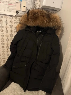 Svart dunjacka med pälsluva - Säljer en svart dunjacka med stor fluffig päls på luvan. Jackan har flera praktiska fickor med knappar och dragkedja framtill. Perfekt för kalla dagar och har en riktigt schysst look med sin mörka färg och rejäla huva.