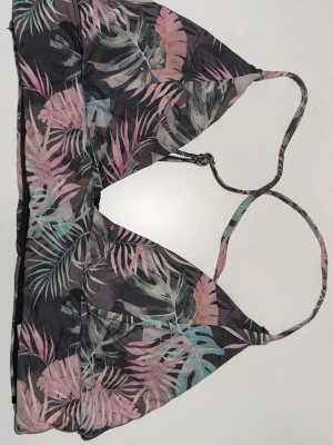 Mönstrad Bikini topp med smala band - Säljer en mönstrad bikini topp  med tropiskt bladmönster i rosa, turkos, grått och svart. Bikini toppen har smala axelband som korsas i ryggen och en omlottliknande front. Materialet känns som syntet och är lätt och luftigt, perfekt för varma dagar.