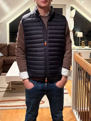 Mörkblå dunväst från Save The Duck - Säljer en svart dunväst från Save The Duck med horisontella quiltade paneler och orange detaljer på dragkedjan. Västen har ståkrage och Save The Duck-logga på nederkanten. Perfekt att slänga över en hoodie eller tröja för extra värme. Västen är sprillans ny och skriv vid minsta fundering🙌🏻🙌🏻