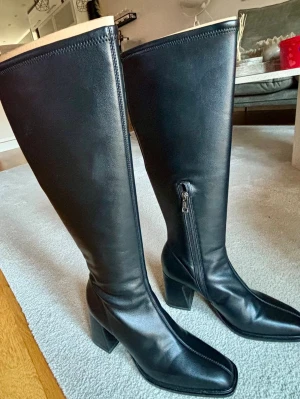 Svarta höga stövlar med blockklack - Säljer ett par svarta höga tamaris stövlar med blockklack och spetsig tå. Tillverkade i 100% imiterat skinn och syntetmaterial. Stövlarna har en elegant design med dragkedja på sidan och passar perfekt till dig som gillar stilrena och trendiga skor. 1800kr ny pris. De är oanvända.