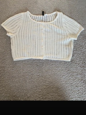 Vit stickad kofta från H&M DIVIDED - Kort, vit stickad kofta från H&M DIVIDED i storlek S. Koftan har ett luftigt hålmönster, rund halsringning och korta ärmar. Stängs med små vita knappar framtill och har en croppad passform som ger en fräsch och trendig look. Jättefint med ett linne under. Skriv om ni har funderingar!❤️☺️