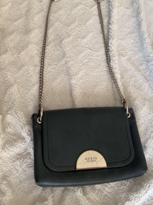 Svart axelväska från Guess - Snygg svart axelväska från Guess med guldfärgad metallplatta framtill och kedjerem. Väskan är tillverkad i skinnimitation och har ett praktiskt fack med dragkedja på insidan. Perfekt storlek för det viktigaste och stilren design.