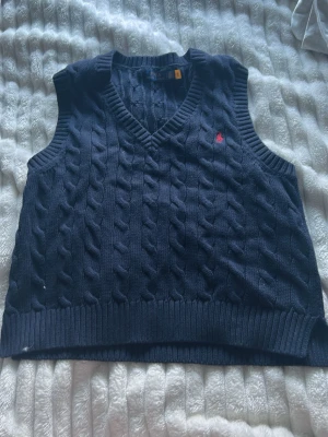 Mörkblå stickad väst från Polo Ralph Lauren - Säljer en mörkblå stickad väst från Polo Ralph Lauren i storlek L. Västen har klassiskt flätstickat mönster, v-ringad hals och en röd broderad logga på bröstet. Ribbad kant vid hals, ärmhål och nederkant. Perfekt för lager-på-lager och snygg till jeans.