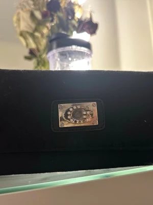 Svart plånbok från Guess med logga - Snygg svart plånbok från Guess med metallplatta och glittrande stenar på framsidan. Plånboken har ett diskret mönster med Guess-loggor, flera fack för kort och mynt, samt dragkedja inuti. Perfekt för dig som gillar stilrena accessoarer med lite bling.