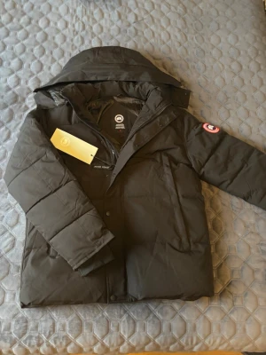 Svart dunjacka från Canada Goose - Svart dunjacka från Canada Goose med huva och klassisk logga på ärmen. Jackan har lång ärm, dragkedja och tryckknappar framtill. Fylld med dun och fjädrar för extra värme, perfekt för kalla dagar. Snygg och stilren design som håller dig varm hela vintern. Jackan är aldrig använd och päls till luvan ingår. Fungerande QR-kod som tar dig direkt till Canada Goose hemsida. Priset kan diskuteras men skambud ignoreras.