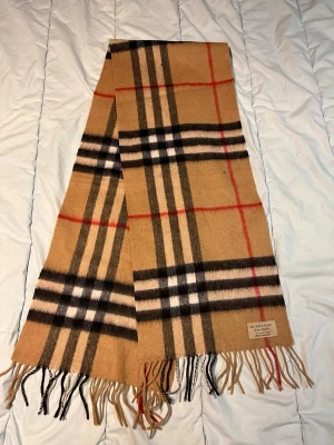 Rutig Burberry halsduk i kashmir - Säljer en klassisk rutig halsduk från Burberry i mjuk 100% kashmir. Färgerna är beige, svart, vit och röd med fransar i ändarna. Den har det ikoniska Burberry-mönstret och en snygg etikett. Perfekt för att lyfta din outfit med lite lyxig London-vibe.