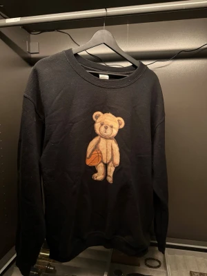 Svart Fashion Bear Tjocktröja    - Svart sweatshirt i storlek M från Fashion Bear, med ett stort tryck av en beige nalle som håller en basketboll på framsidan. Tröjan har rund hals och långa ärmar, tillverkad i en mjuk bomullsblandning som är perfekt för chill dagar.