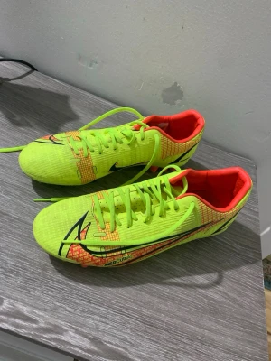 Nike Mercurial fotbollsskor neon - Säljer ett par Nike Mercurial fotbollsskor i neonljus gul med orange insida och svarta detaljer. Skorna har snörning och en platt sula, designade för att ge bra grepp på planen. Materialet är syntet och skon har en rund tå. Perfekt för dig som vill sticka ut på fotbollsplanen.