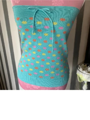 Turkos prickig bandeau tubtopp - Säljer en turkos bandeau tubtopp med färgglada prickar i rosa, gul och orange. Toppen har en söt knytning framtill och är stickad i mjukt syntetmaterial. Perfekt för sommarens alla häng och ger en riktigt glad vibe.