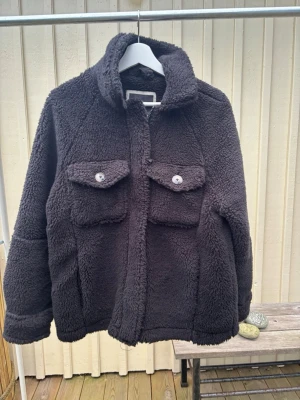 Svart teddyjacka  - Svart teddyjacka från FB Sister i storlek S. Jackan har två stora bröstfickor med knappar, hög krage och är helfodrad i fluffigt teddy-material. Perfekt för kalla dagar när du vill vara både varm och snygg.