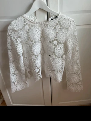 Vit spetsblus med lång ärm - Säljer en vit blus med lång ärm i vacker spets med blommigt mönster. Blusen har rund hals och är genombruten med detaljerade virkade blommor över hela plagget. Perfekt för dig som gillar romantisk och feminin stil.