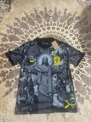 Nike Brasil t-shirt med grafiskt tryck - Snygg svart Nike t-shirt med Brasil-logga och unikt grafiskt tryck framtill som visar Kristusstatyn och brasilianska symboler. Gula detaljer och flagga på ärmen. Perfekt för dig som gillar fotboll och streetwear.
