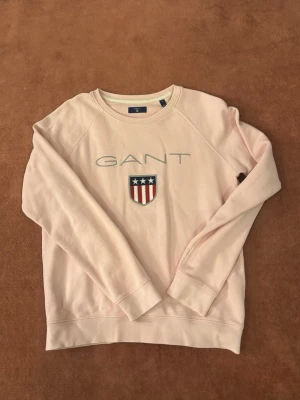 Ljusrosa sweatshirt från GANT - Säljer en ljusrosa sweatshirt från GANT i storlek S. Tröjan har ett broderat GANT-logo och ett emblem med stjärnor och ränder på bröstet. Rund halsringning och långa ärmar, tillverkad i mjuk bomull för skön komfort.
