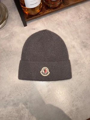 Grå Moncler stickad mössa - Snygg grå stickad mössa från Moncler med klassisk ribbad design och uppvikt kant. Mössan har Moncler-logga framtill i vitt och rött. Perfekt för att hålla värmen och addera lite lyx till din vinterstil.