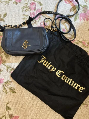 Svart axelväska från Juicy Couture - Säljer en svart axelväska från Juicy Couture i skinn med guldfärgade detaljer och kedja. Insidan har ett blommigt mönster i grönt, rött och blått. Väskan har magnetlås och JC-logga framtill samt en dekorativ JC-berlock på sidan. Perfekt för dig som gillar statement-accessoarer.