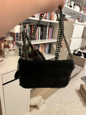 stella mccartney liknande - Säljer en svart axelväska i mocka-look med grov kedjerem i metall. Väskan har en rektangulär form och dekorativa kedjedetaljer runt kanterna. Baksidan har en liten ficka med dragkedja. Perfekt för dig som gillar en edgy stil.