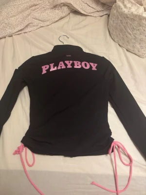  Playboy kofta - Svart kofta från Playboy med dragkedja framtill, rosa logga på bröstet och stor rosa PLAYBOY-text på ryggen. Långärmad med rosa snörning i sidorna och två fickor med dragkedja. Snygg och trendig design som sticker ut. Från misguided.