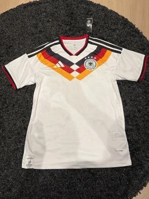 Vit Adidas fotbollströja Tyskland - Snygg vit fotbollströja från Adidas med Tysklands landslagsmärke och coolt geometriskt mönster i svart, rött och gult över bröstet. Tröjan har svarta och vita ränder på axlarna, röd kant vid hals och ärmslut samt Climacool-material som andas.