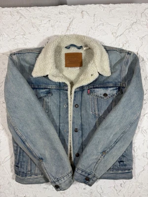 Levi's jeansjacka med teddyfoder - Säljer en Levi's Premium jeansjacka i ljusblå denim med mjukt vitt teddyfoder. Jackan har klassisk krage, bröstfickor med knappar och knappstängning framtill. Perfekt för dig som vill ha en chill och varm look under höst och vinter.