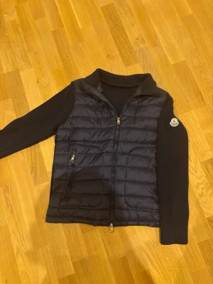 Marin blå cardigan från Moncler  - Säljer en Marin blå cardigan från Moncler med quiltad front och två dragkedjefickor. Cardiganenhar ribbstickade partier på sidor och krage samt en tydlig Moncler-logga på axeln. Perfekt för lager-på-lager och riktigt snygg till höst och vinter.