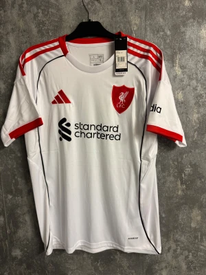 Vit Liverpool tröja - Ny storlek L - Bilderna visar en vit och röd Liverpool FC fotbollströja från Adidas, inte skor. Tröjan har röda detaljer och klubbmärke på bröstet.